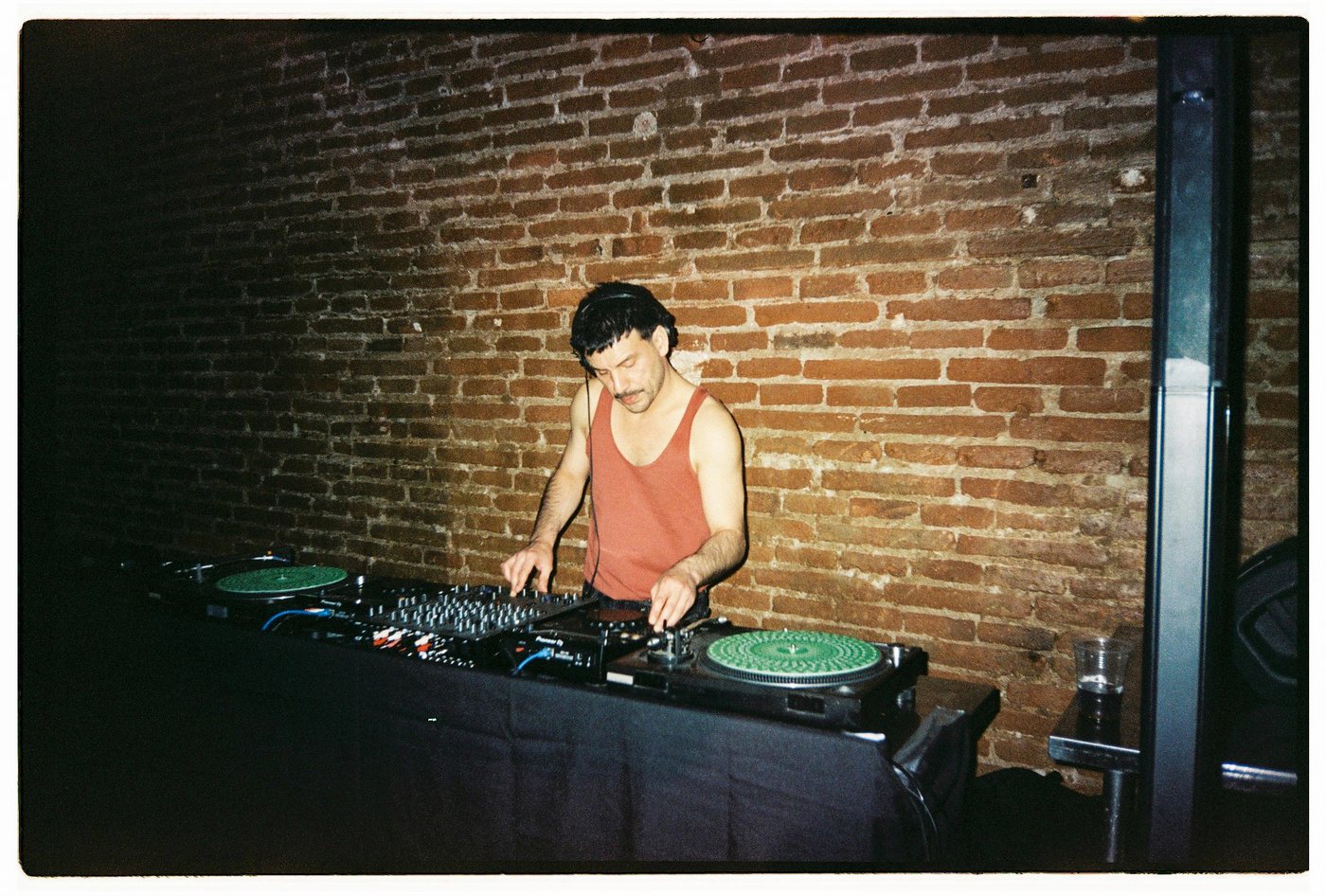DJ in a red tank top at turntables before a brick wall