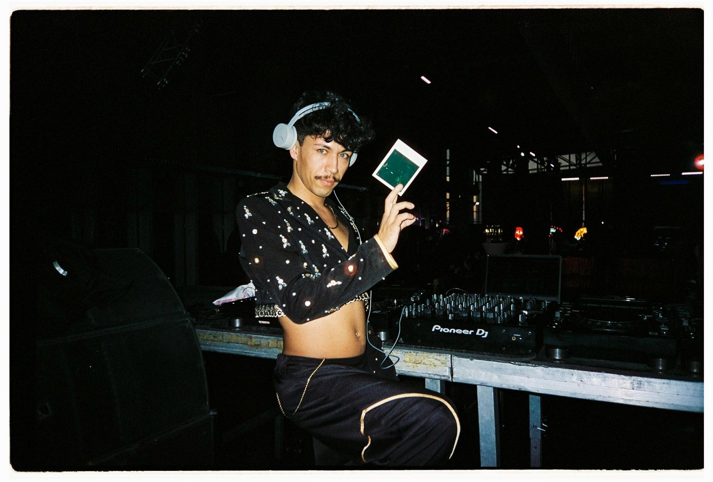 DJ in a sequin jacket and headphones holding up a Polaroid
