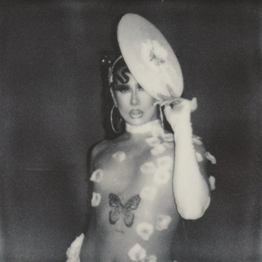 Person in a sheer floral bodysuit holding a large hat to their head